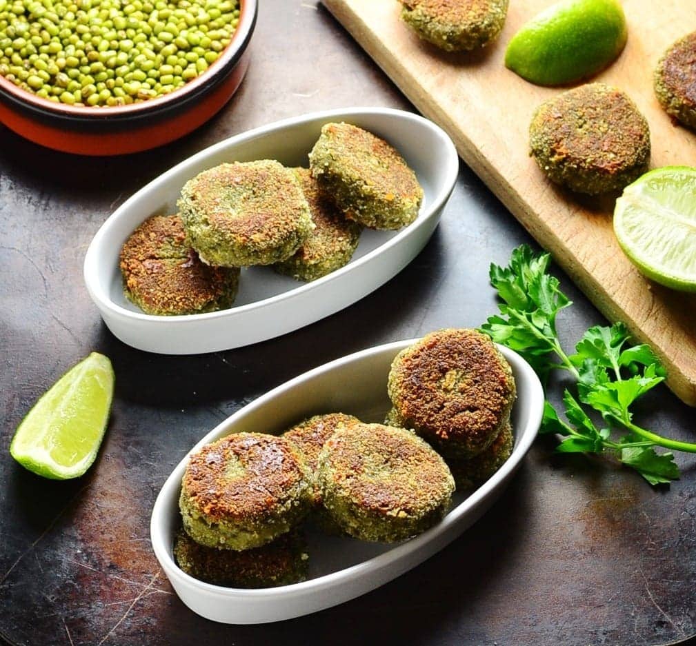 Side view of bean patties in white oval dishes with lime, beans in brown dish and cutting board on oven tray.