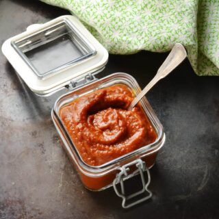 Barbecue sauce in square jar with spoon and green cloth at the back on, on dark brown surface.