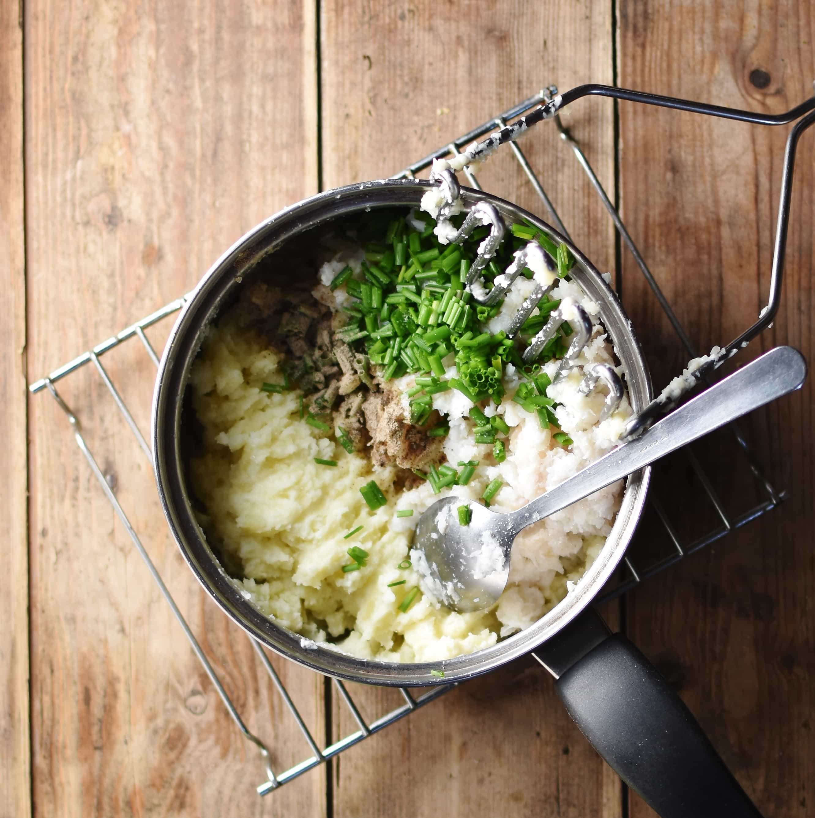 Fish cakes ingredients in pot with spoon and potato masher, on top of cooling rack.