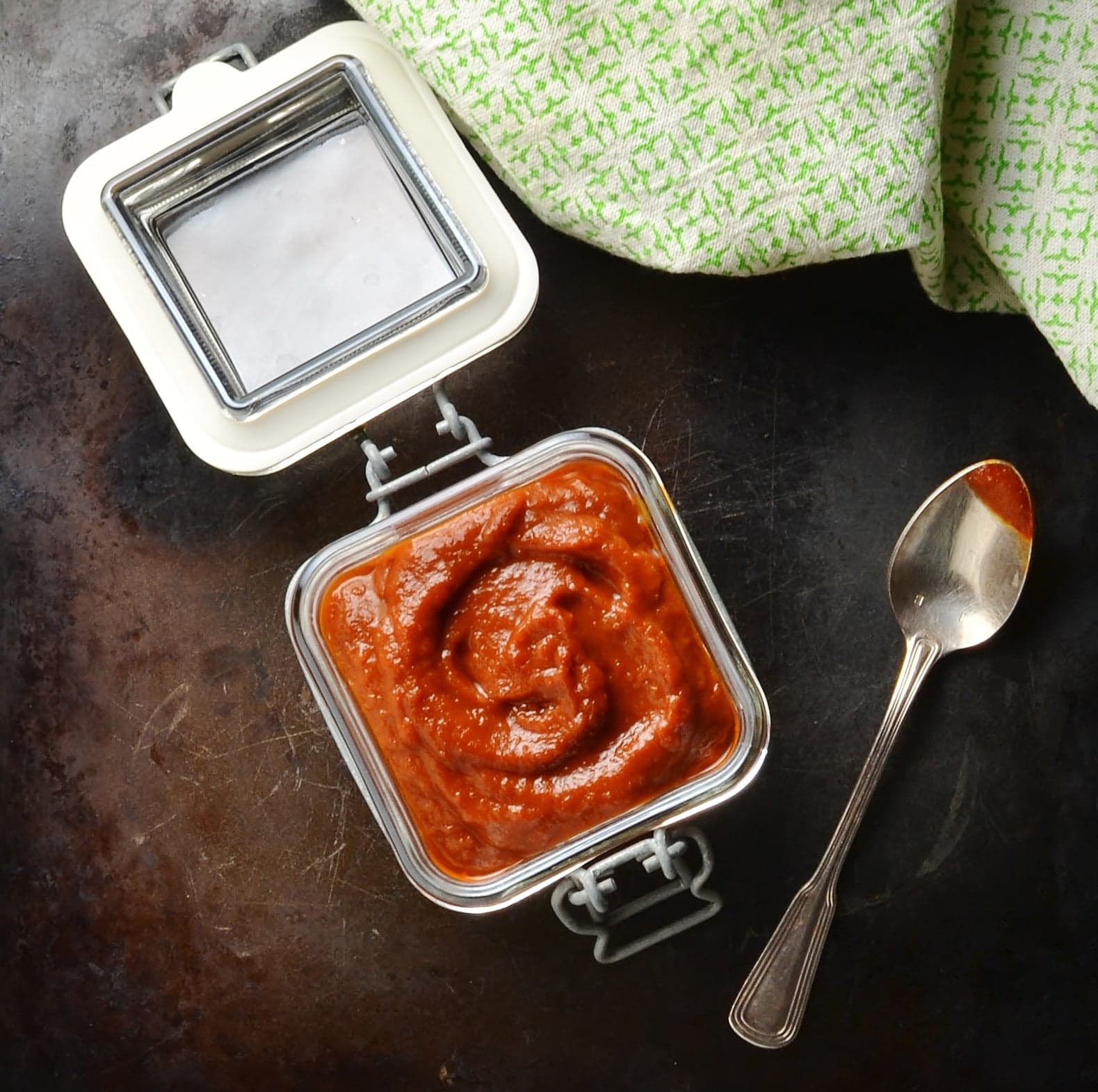 Barbecue sauce in open jar with spoon and green cloth on dark brown surface.