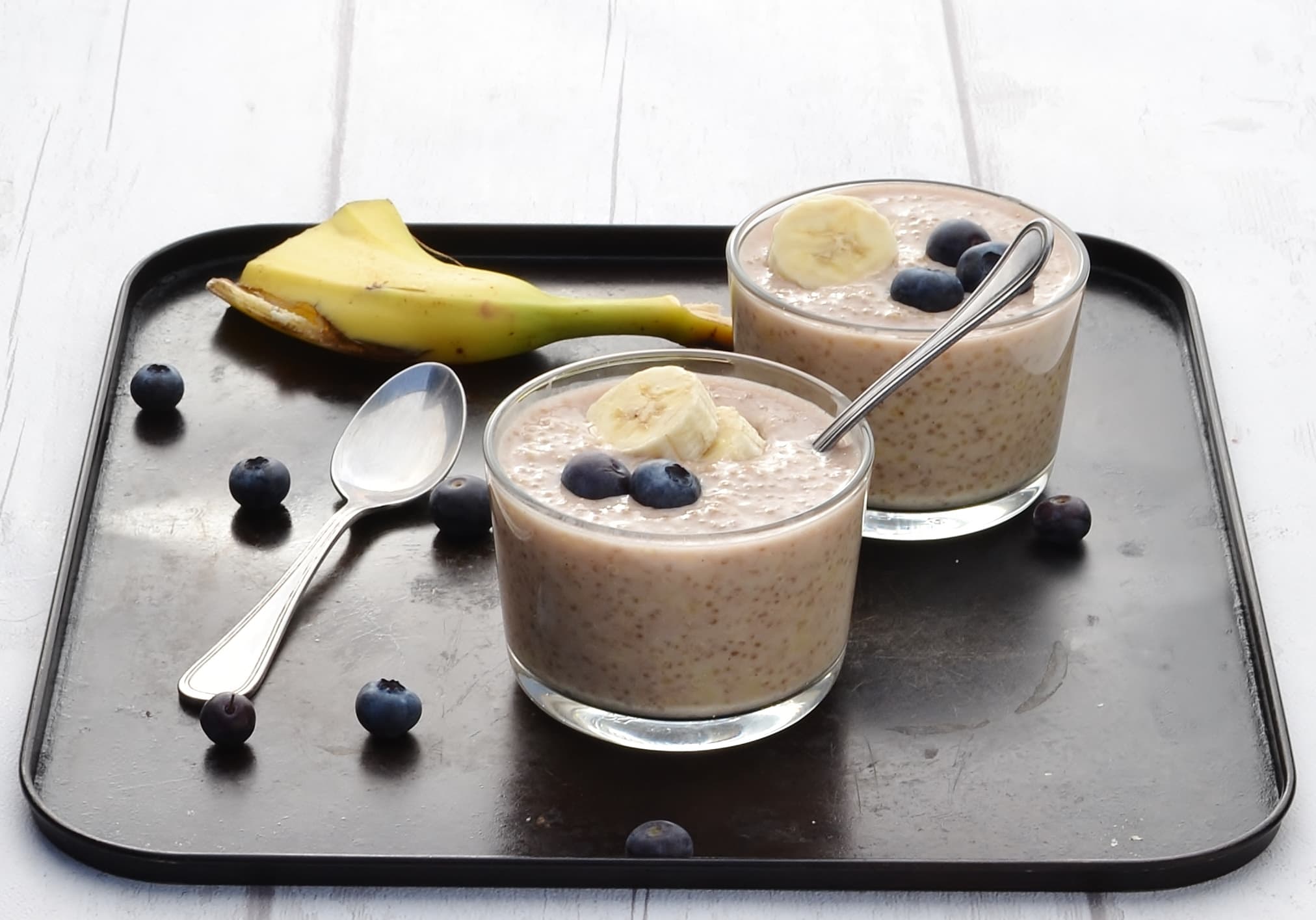 Side view of overnight quinoa with banana slices and blueberries in 2 cups with spoon, another spoon, blueberries and banana peel on dark table.