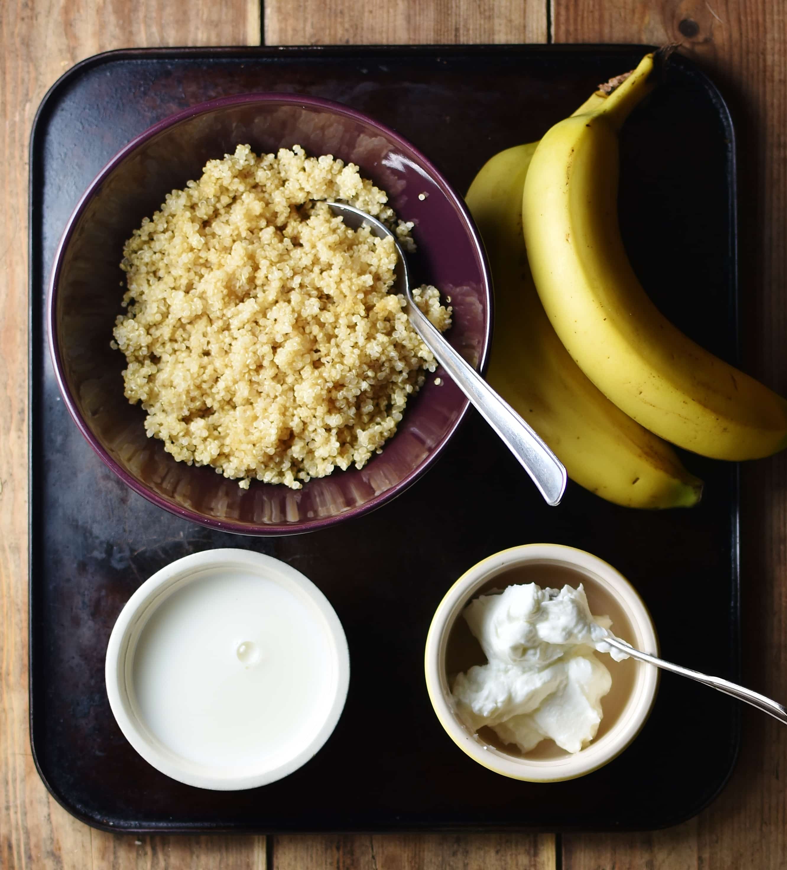 Cooked quinoa in purple bowl with spoon, 2 bananas, yogurt in small dish with spoon and milk in another dish.