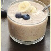 Side view of overnight quinoa with blueberries and banana slices and spoon in see-through glass, banana and another glass in background.