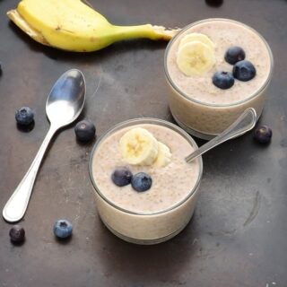 Top down view of overnight quinoa with banana slices and blueberries in 2 cups with spoon, another spoon, blueberries and banana peel on dark table.