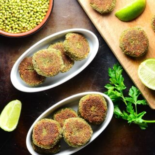 Top down view of mung bean mushroom patties in white oval dishes with lime, beans in brown dish and cutting board on oven tray.