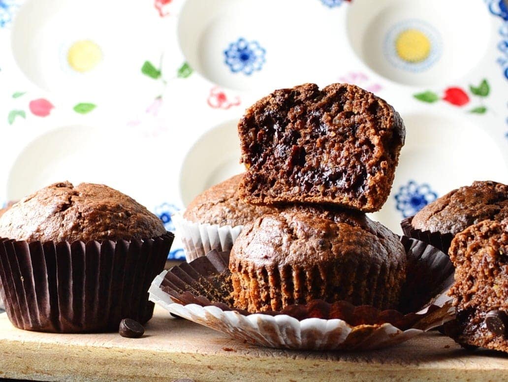 Side view of chocolate muffins with paper cases on wooden board with ceramic muffin dish in background.