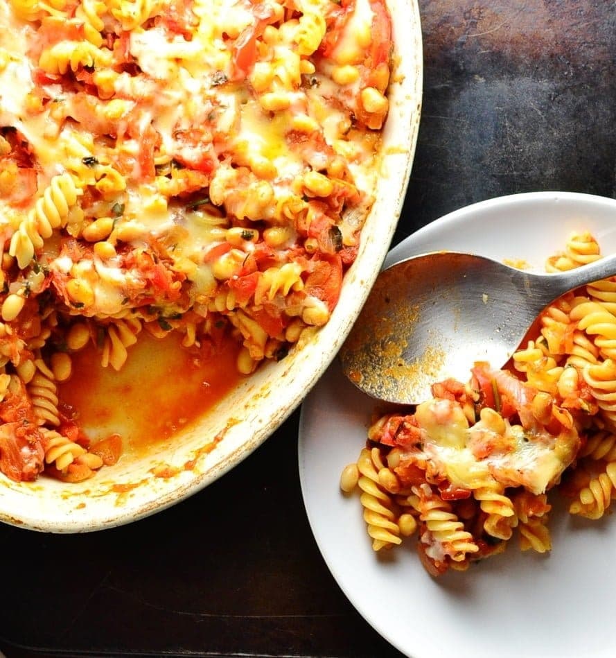 Top down view of bean pasta bake in oval casserole dish and pasta on white plate with spoon on oven tray.