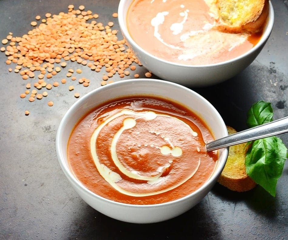 Side view of tomato and lentil soup in 2 white bowls with spoon, toasted bread, lentils and basil leaves on oven tray.  