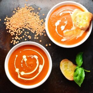 Top down view of tomato and lentil soup in 2 white bowls with toasted bread, lentils and basil leaves on oven tray.  