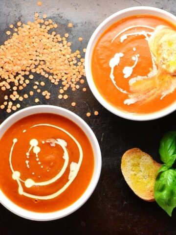 Top down view of tomato and lentil soup in 2 white bowls with toasted bread, lentils and basil leaves on oven tray.  