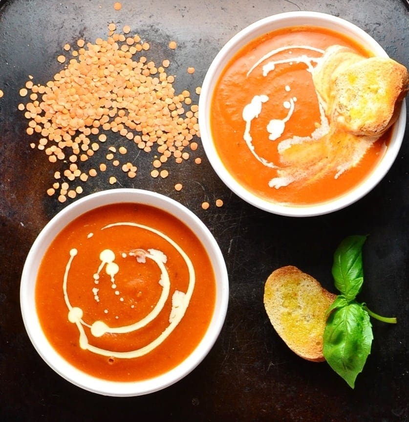 Top down view of tomato soup in 2 white bowls with toasted bread, lentils and basil leaves on oven tray.  