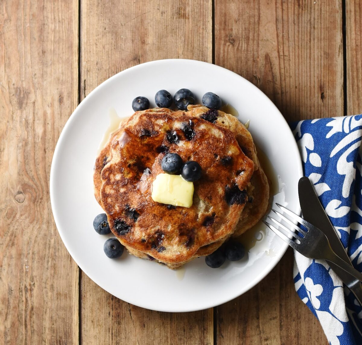 Stack of blueberry pancakes on white plate with blue-and-white cloth with fork and knife to the right.