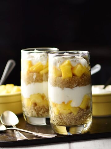 Side view of yogurt, mango, quinoa parfait in 2 tall glasses, 2 spoons and 2 yellow dishes with spoons in background on top of dark tray.