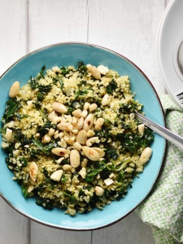 Top down view of bulgur wheat with kale and almonds in blue bowl with spoon, with green cloth and plate with forks and capers to right.