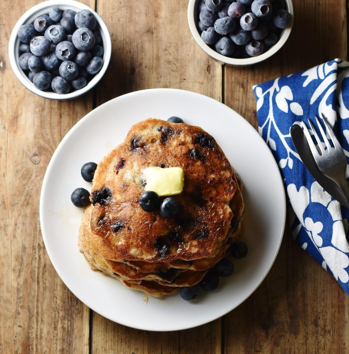 Blueberry pancakes stacked on top of white plate, with blueberries in 2 dishes and blue-and-white patterned cloth in background.
