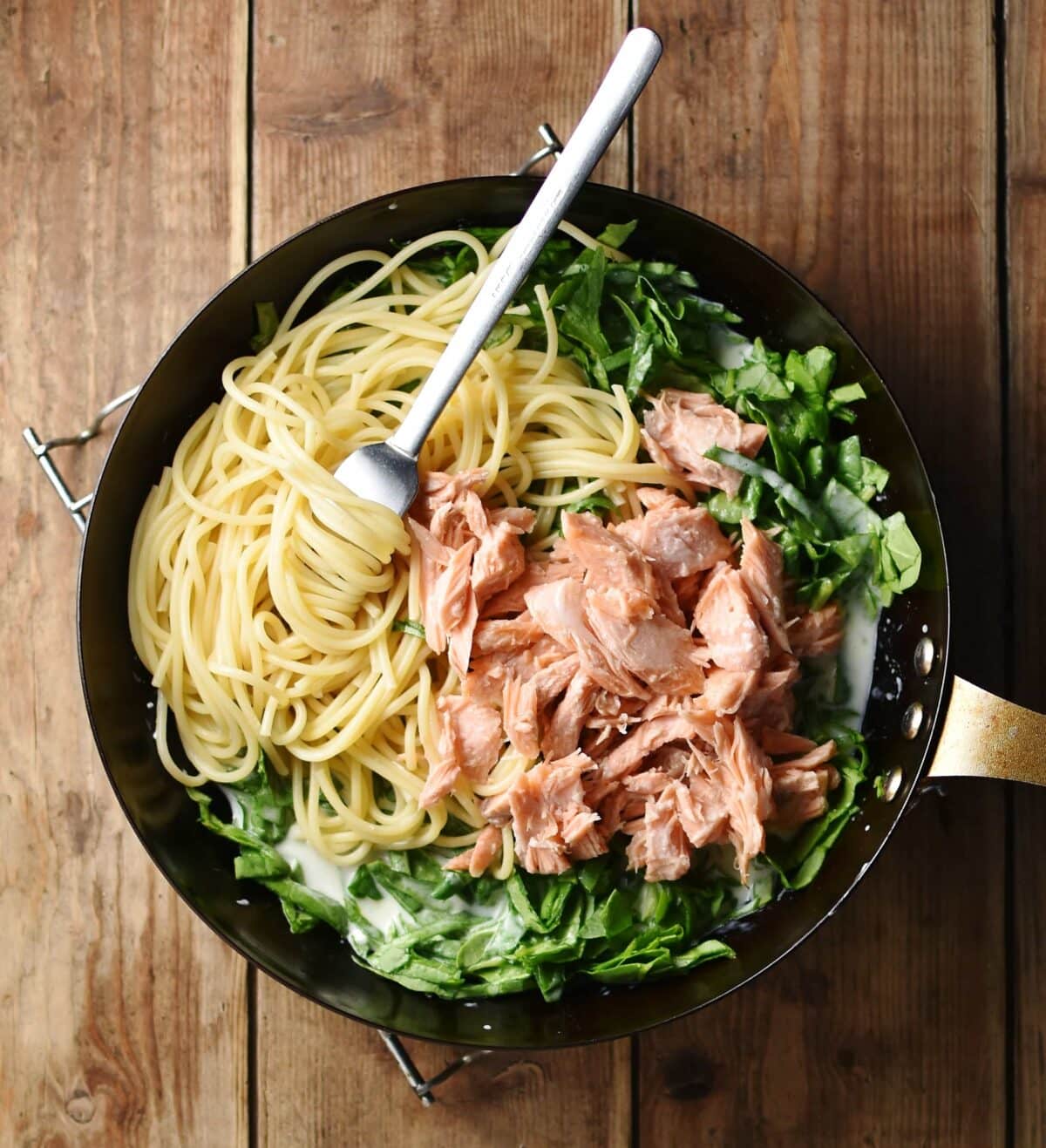 Spaghetti with fork, shredded salmon and chopped spinach in white sauce in large pan.
