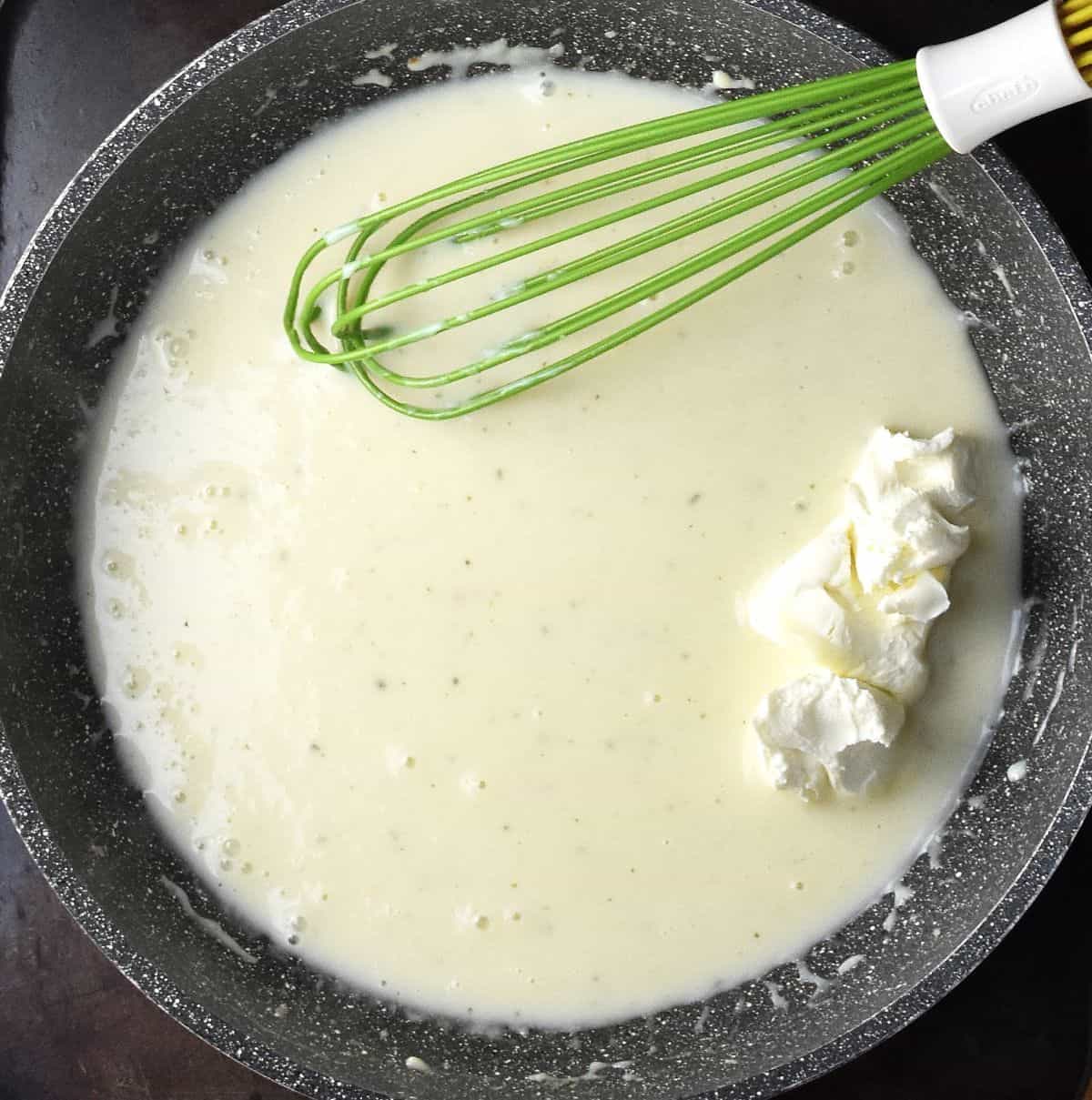 Top down view of creamy sauce with cream cheese in large pan with green whisk.