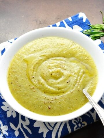 Top down view of cream of zucchini soup in white bowl with spoon on blue cloth.