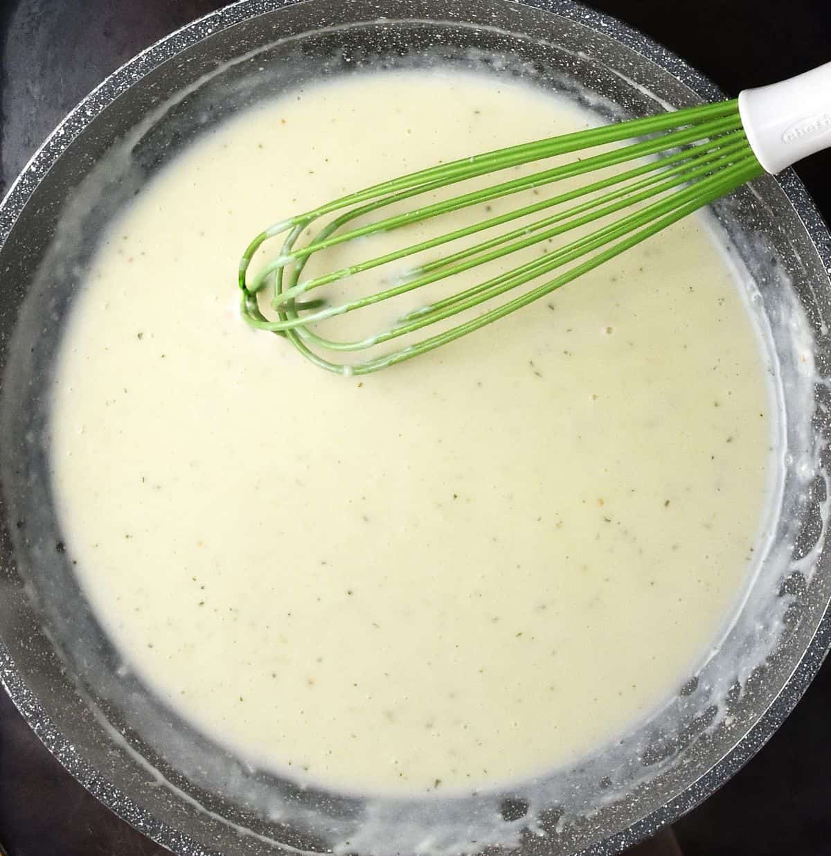 Top down view of creamy sauce in large pan with green whisk.