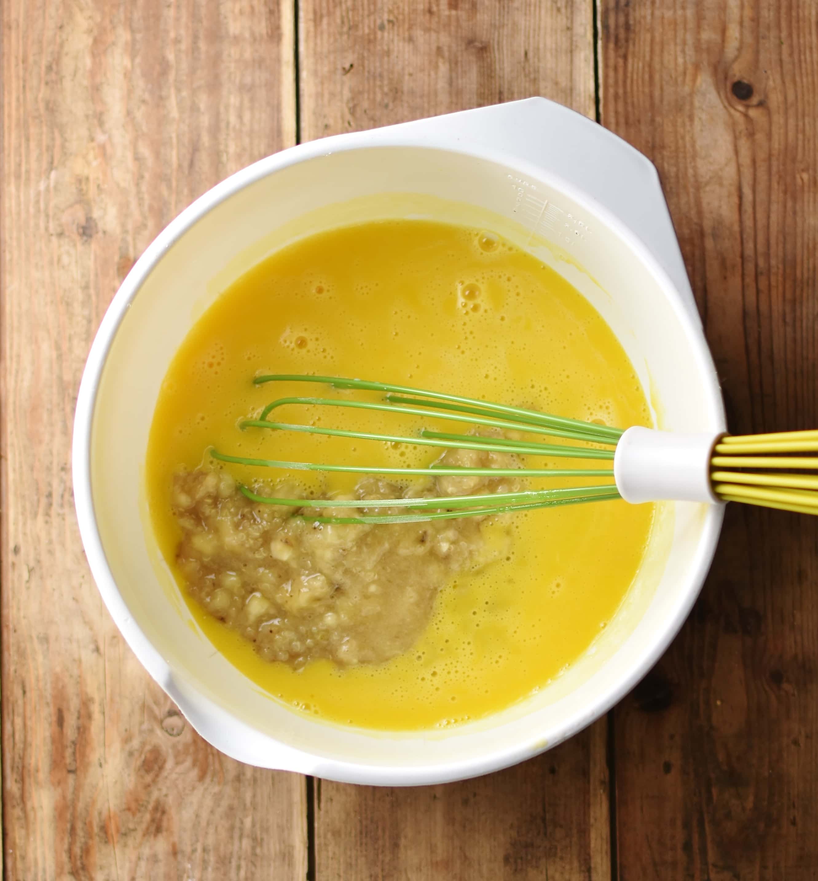 Egg mixture with mashed banana and green whisk in large white bowl.