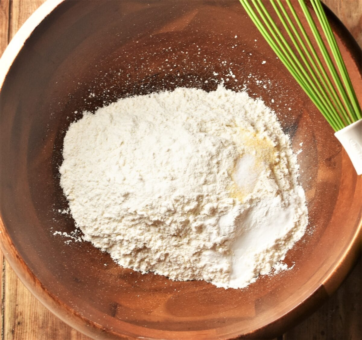 Dry ingredients in wooden bowl with green whisk to the right.