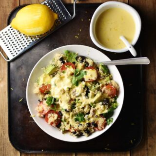 Quinoa vegetable salad in white bowl with spoon, creamy dressing in white dish with spoon in top right and zester with lemon in top left corners.