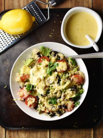 Quinoa vegetable salad in white bowl with spoon, creamy dressing in white dish with spoon in top right and zester with lemon in top left corners.