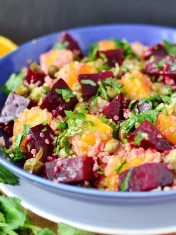 Side view of beets quinoa salad in blue bowl on white plate, with herbs and oranges in background.