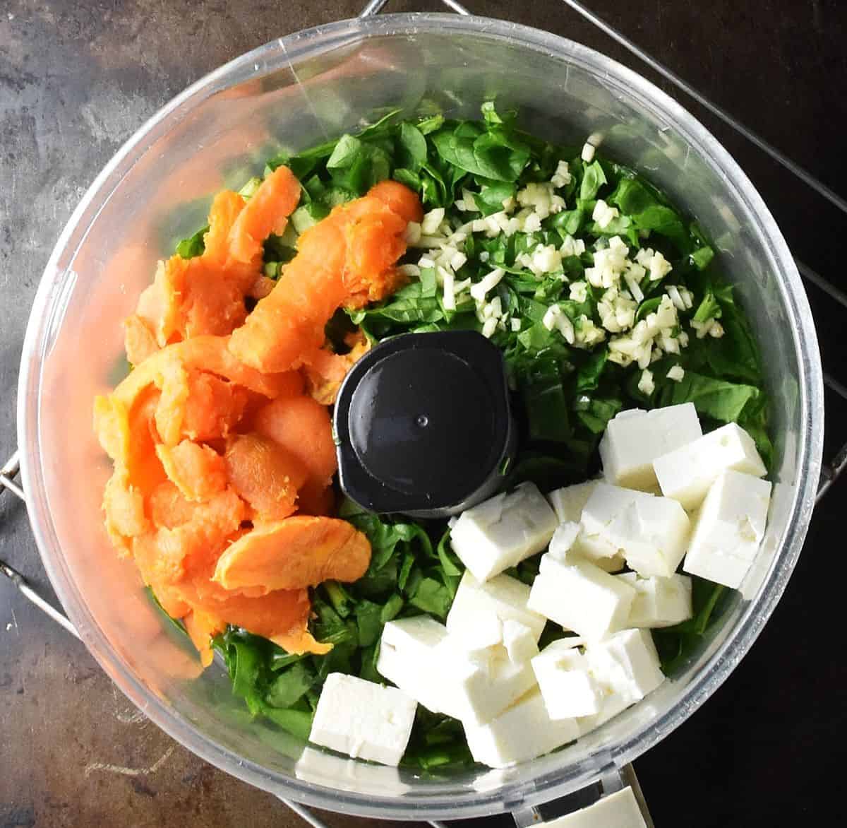 Top down view of sweet potato pulp, chopped spinach and garlic and cubed feta in food processor bowl.