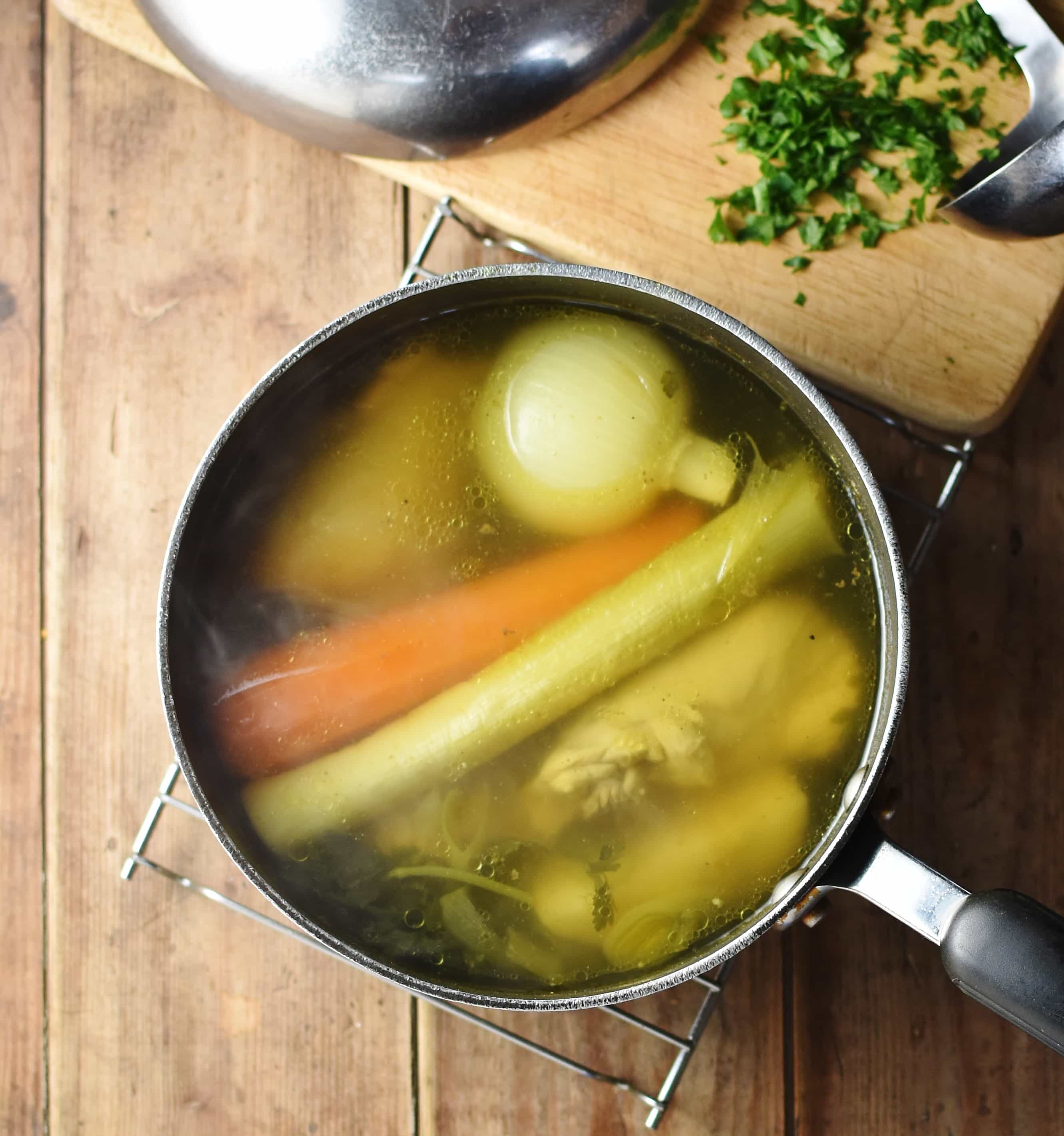 Chicken broth with cooked whole vegetables and chicken pieces in large pot, with chopped herbs and lid on cutting board at the top.