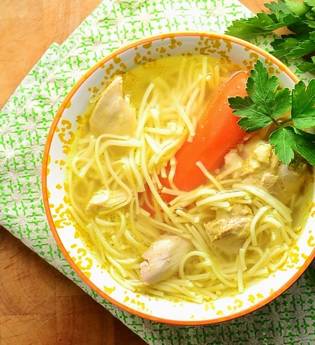 Top down view of chicken noodle soup with carrot and parsley inside yellow-and-white bowl, on top of green cloth.