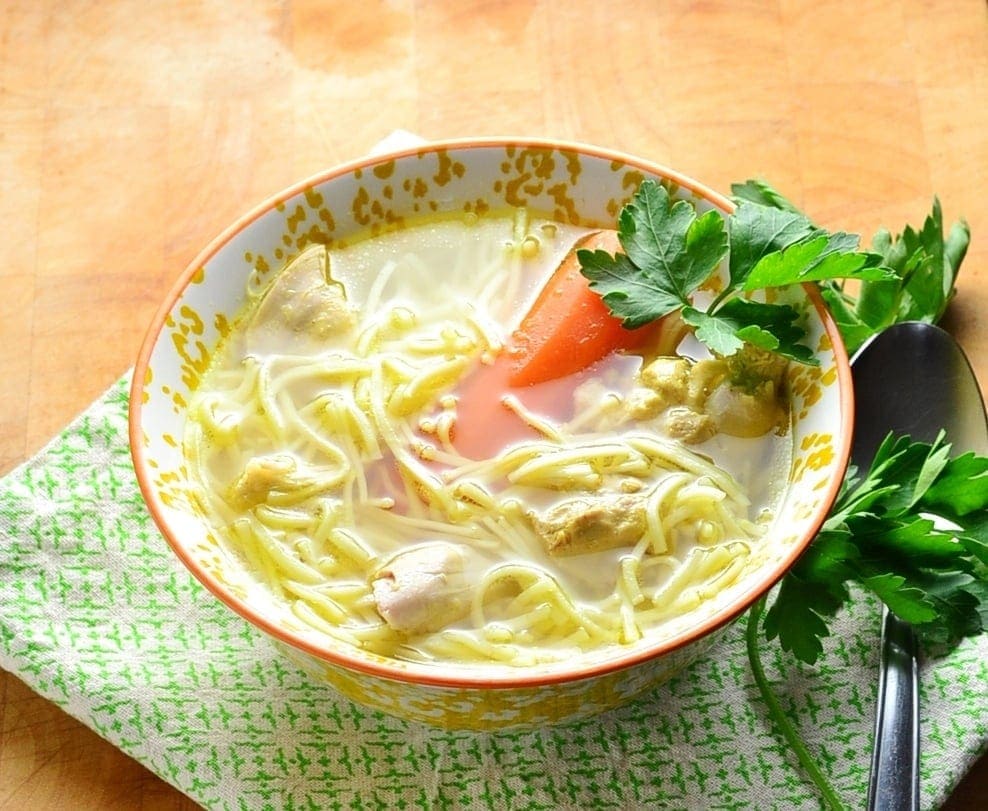 Chicken noodle soup in orange bowl with spoon and parsley on green cloth.