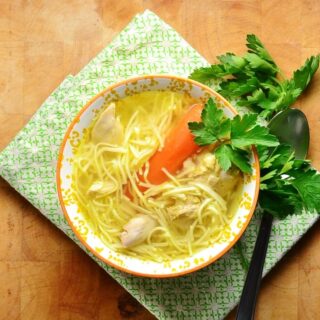 Chicken noodle rosol soup in orange bowl with spoon and parsley on green cloth.