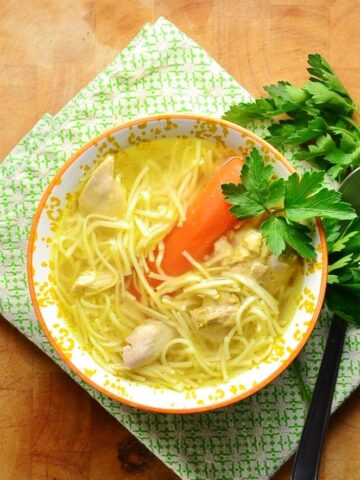 Chicken noodle rosol soup in orange bowl with spoon and parsley on green cloth.