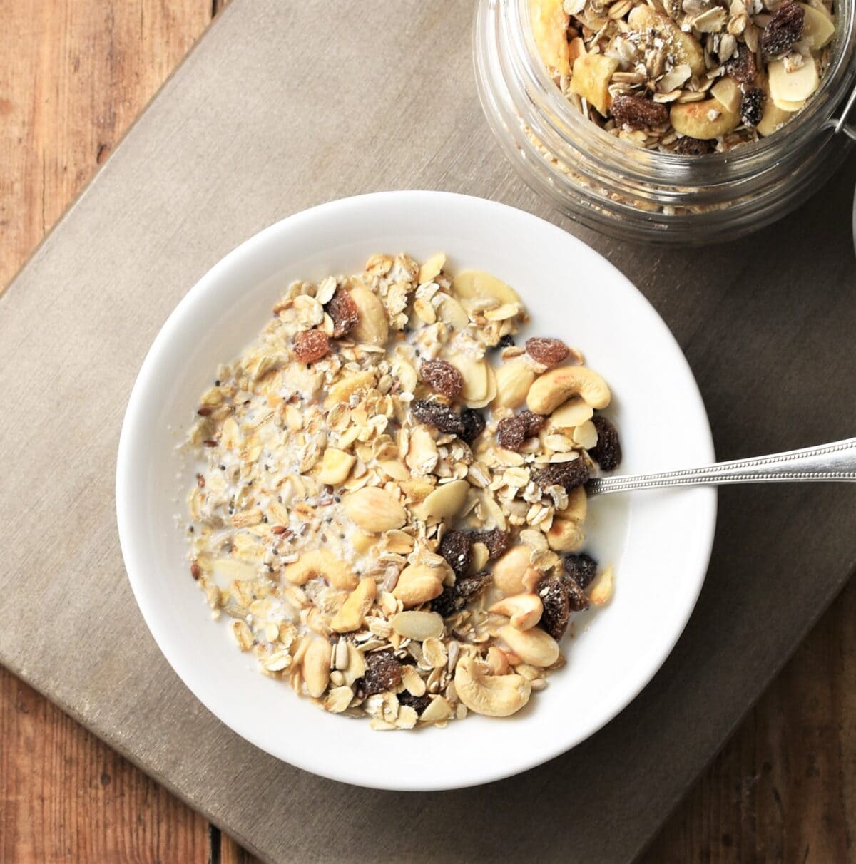 Muesli in white bowl with spoon, with muesli in open jar in top right.