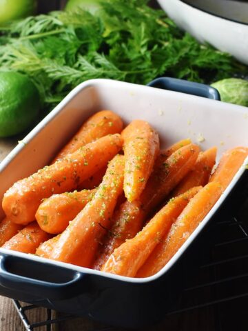 Side view of braised carrots in rectangular blue dish, with limes, carrot tops and white pan in background.