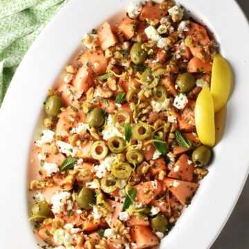 Top down view of cantaloupe salad with feta, olives and lemon wedges on white oval platter.