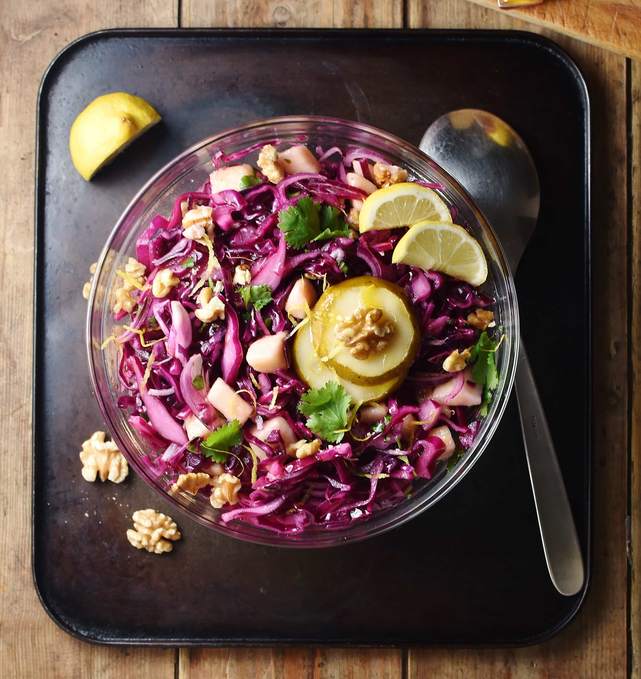 Chopped red cabbage, pear slices, walnuts and herbs in bowl, with large spoon to the right, walnuts and lemon wedge to the left.