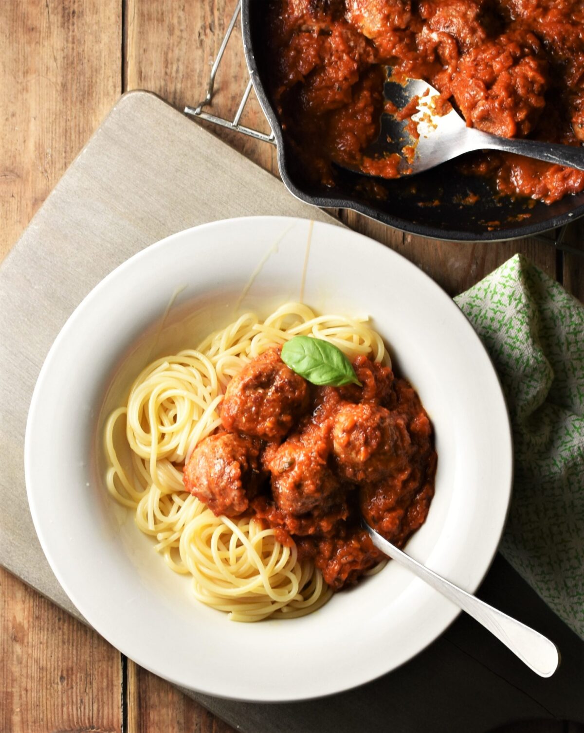 Meatballs in tomato sauce over spaghetti in bowl with fork and meatballs in sauce in pan in top right corner.