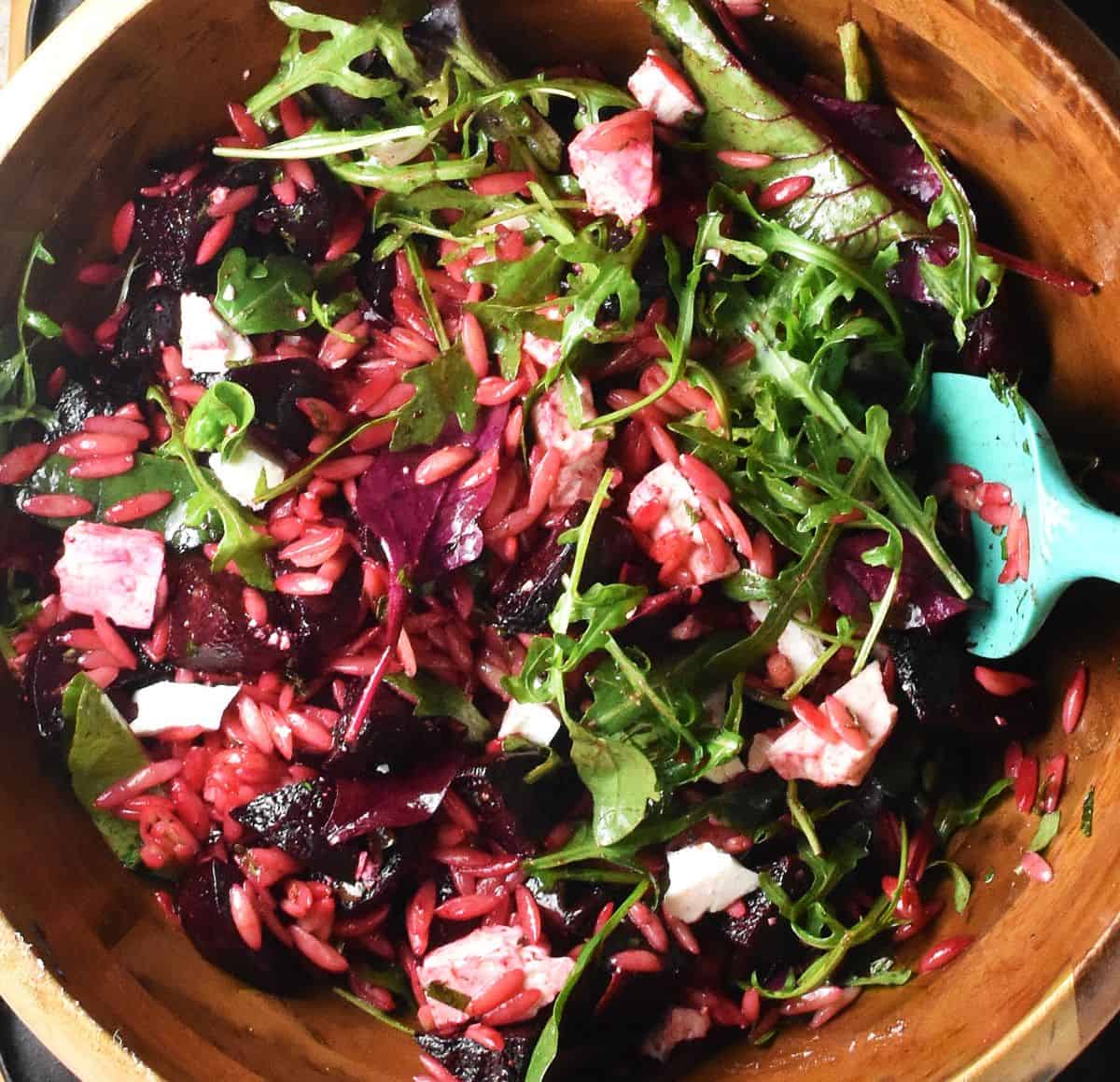 Pasta salad with roasted beet pieces, crumbled feta and salad leaves in wooden bowl with blue spatula.