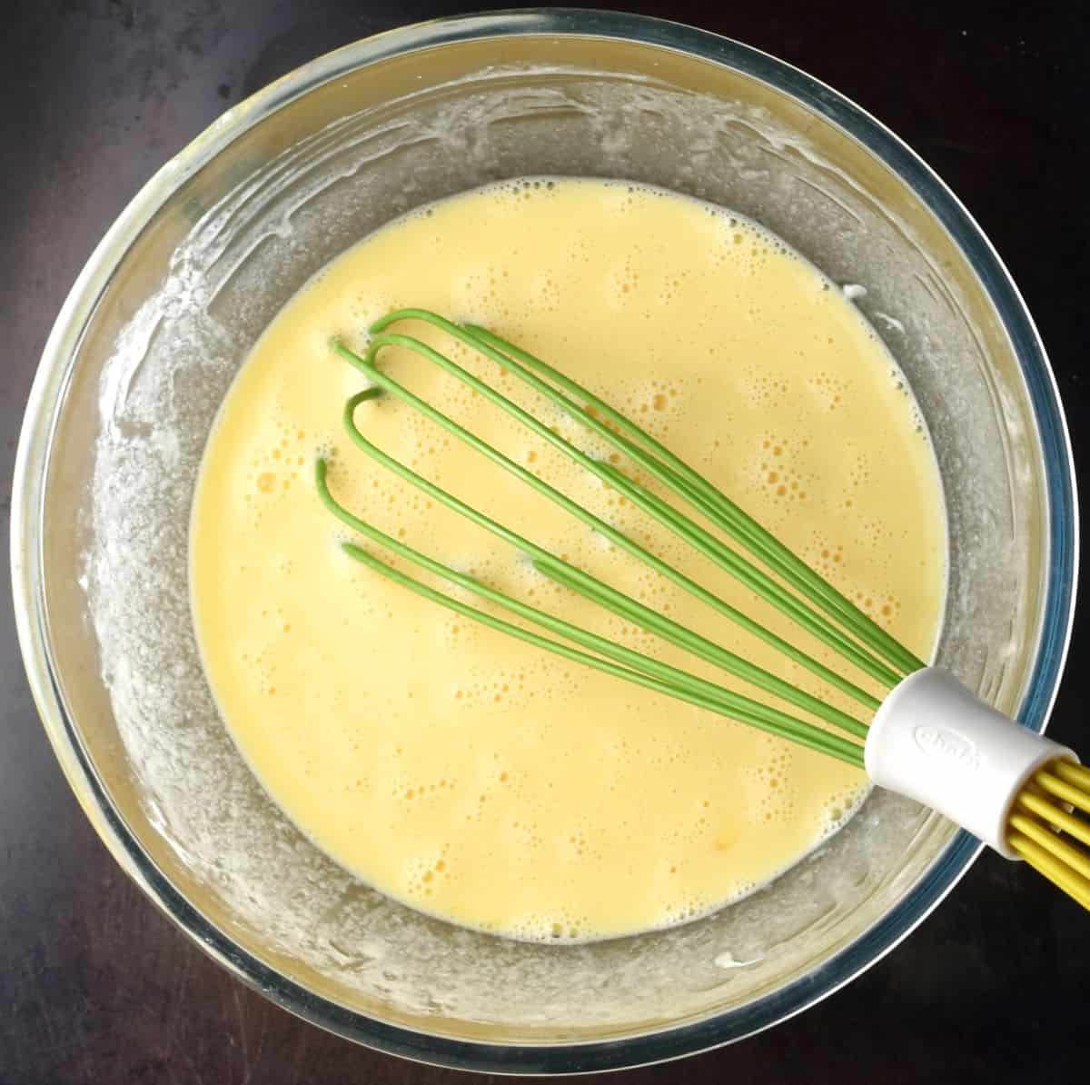 Top down view of smooth egg mixture in bowl with whisk.