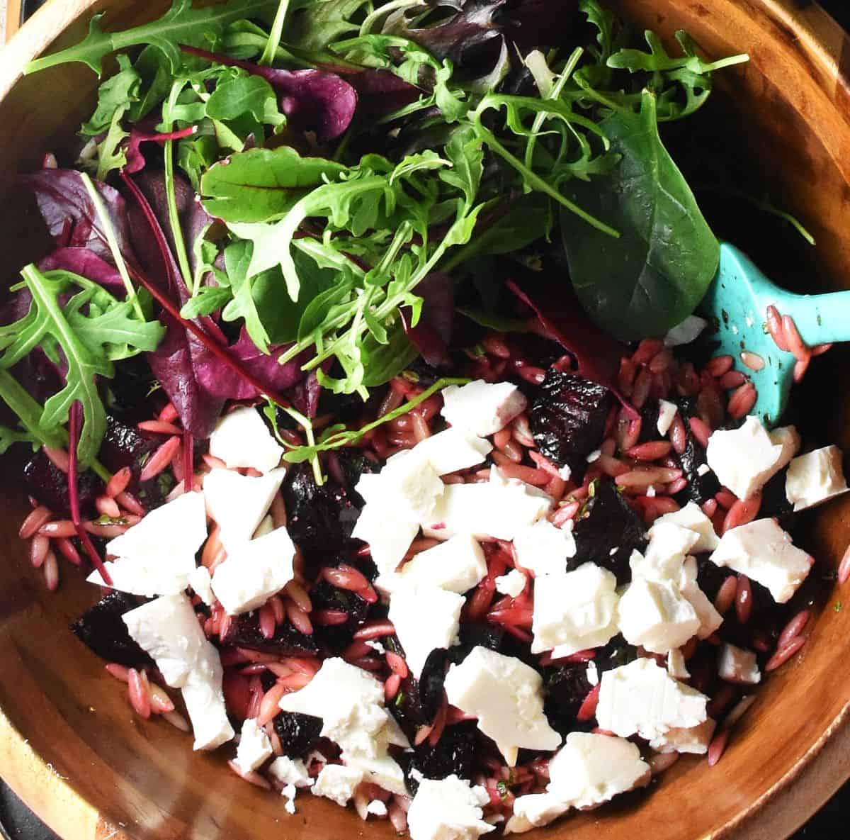 Beet and pasta salad mixture with feta chunks and salad leaves in wooden bowl.