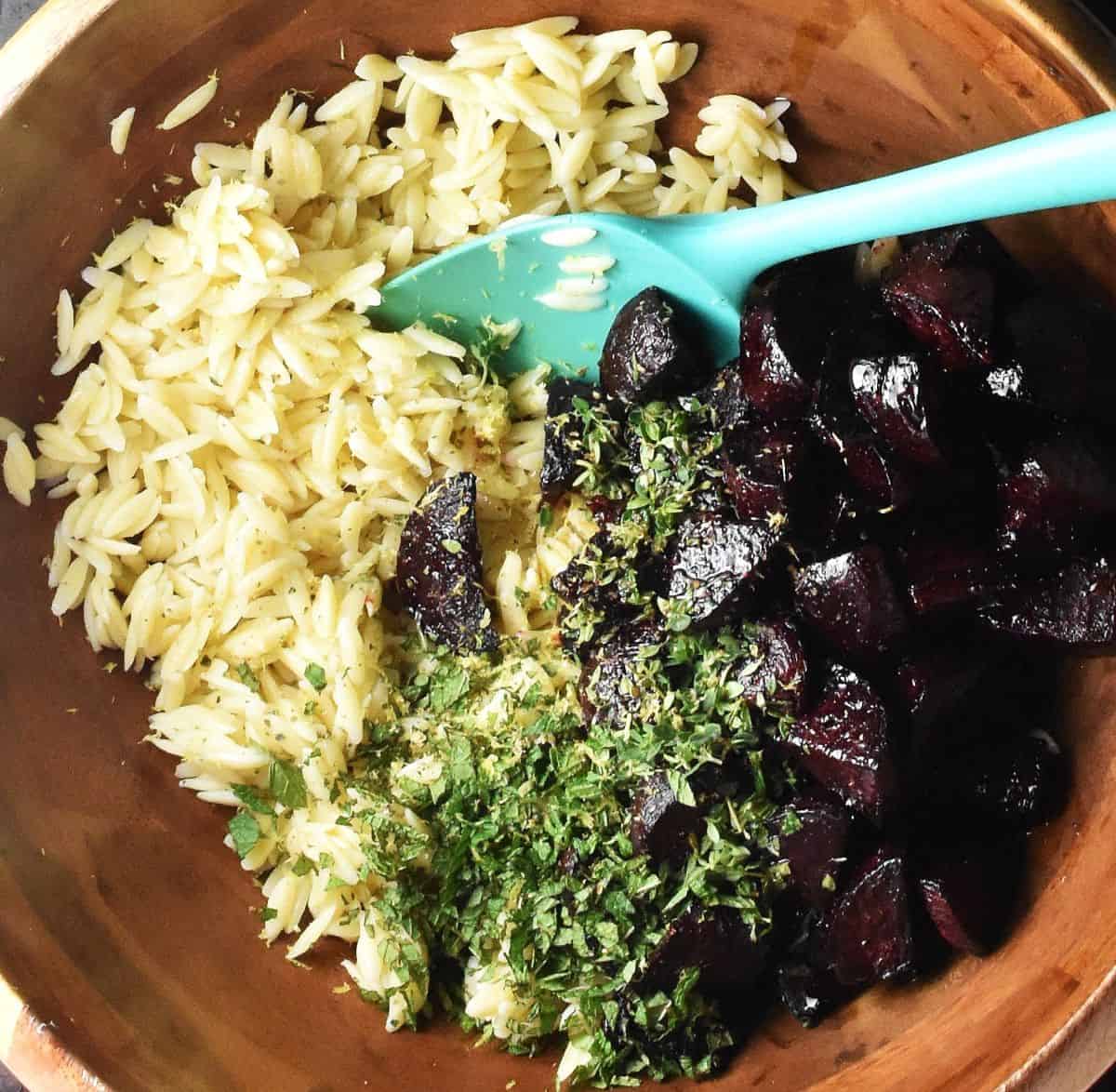Cooked orzo pasta, roasted beet pieces and chopped herbs in wooden bowl with blue spatula.