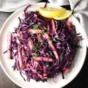 Top down view of red cabbage apple slaw with lemon wedges on pink plate.