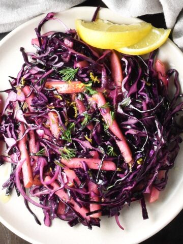Top down view of red cabbage apple slaw with lemon wedges on pink plate.