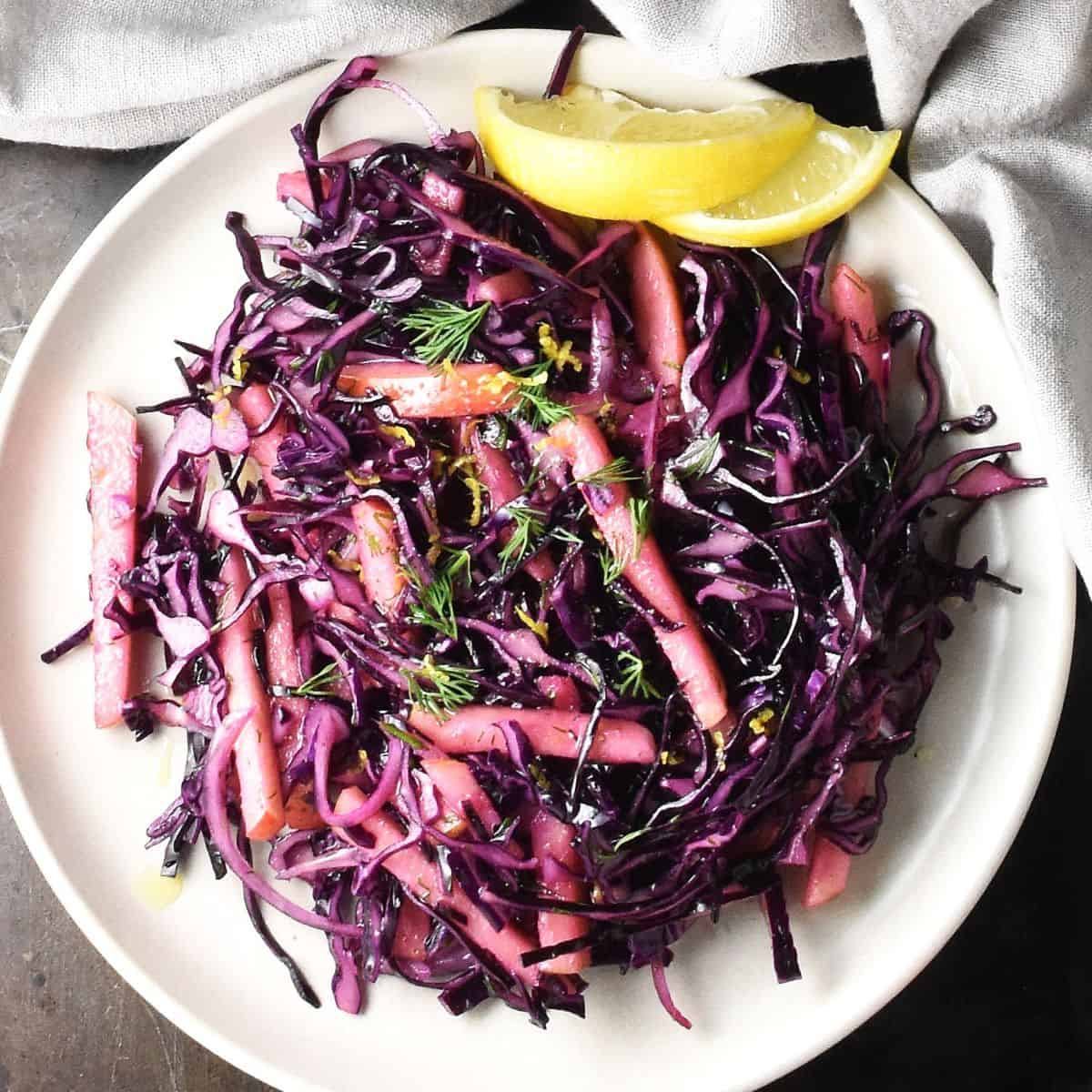 Top down view of red cabbage apple slaw with lemon wedges on pink plate.