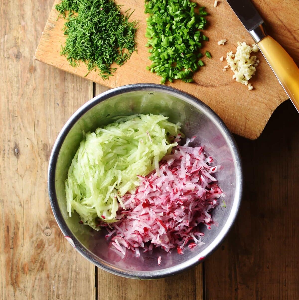 Grated cucumber and radish in metal bowl as well as chopped herbs and garlic on top of cutting board with knife.