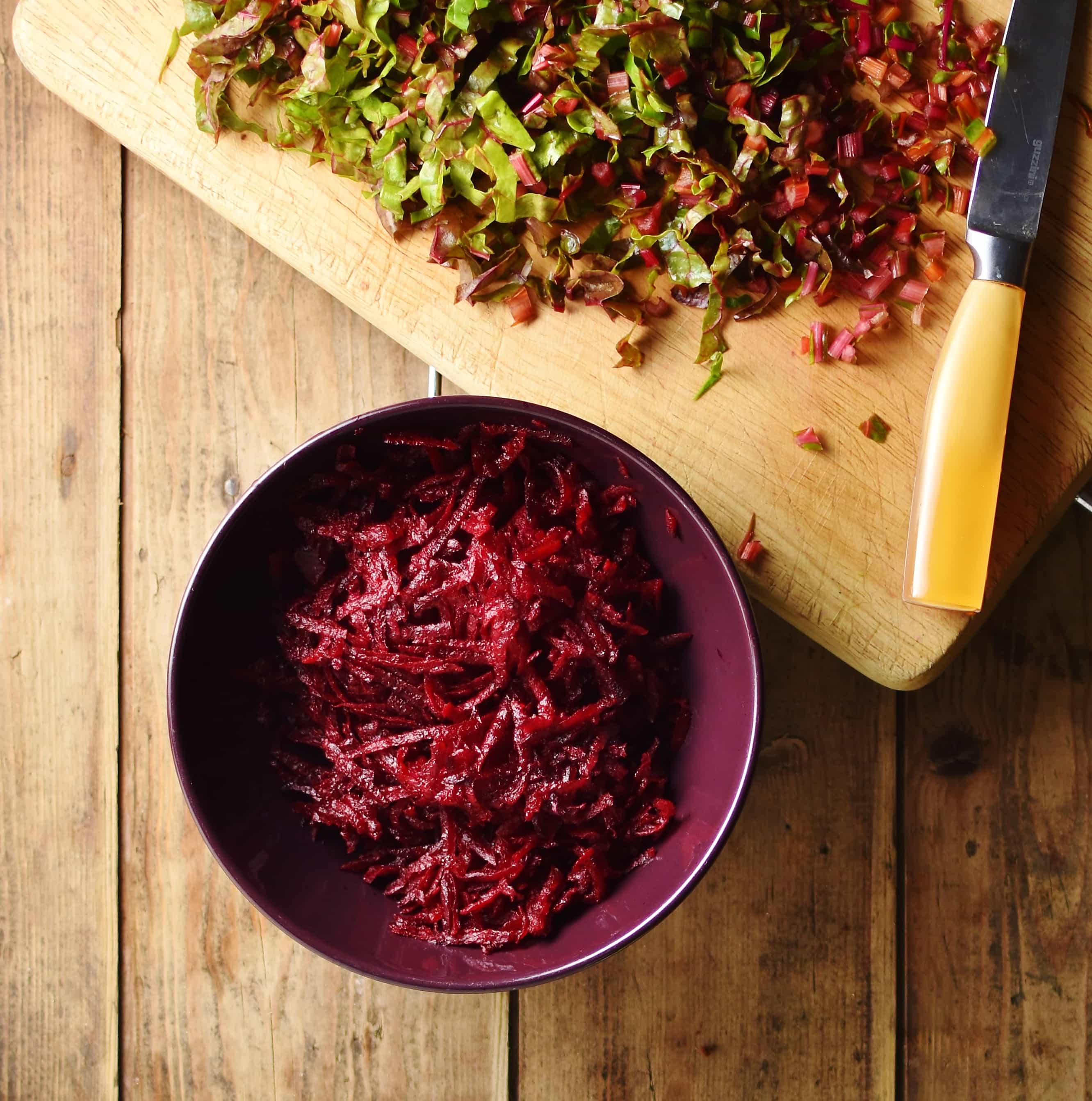 Grated beetroot in purple bowl and chopped beet leaves and knife on top of cutting board.