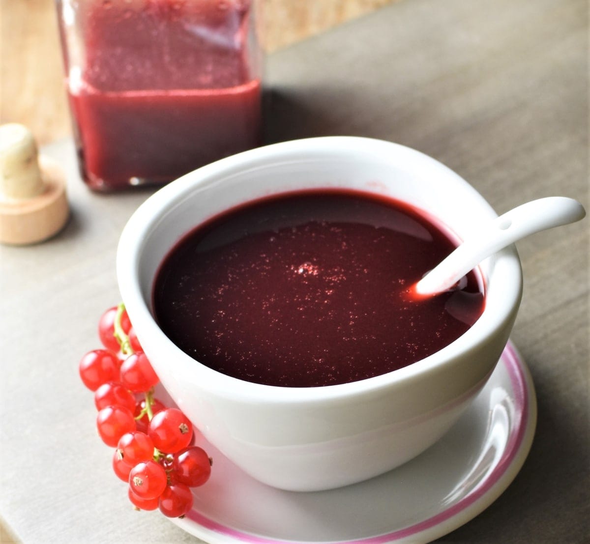 Redcurrant gravy in white bowl with spoon and redcurrants and sauce in bottle in background.