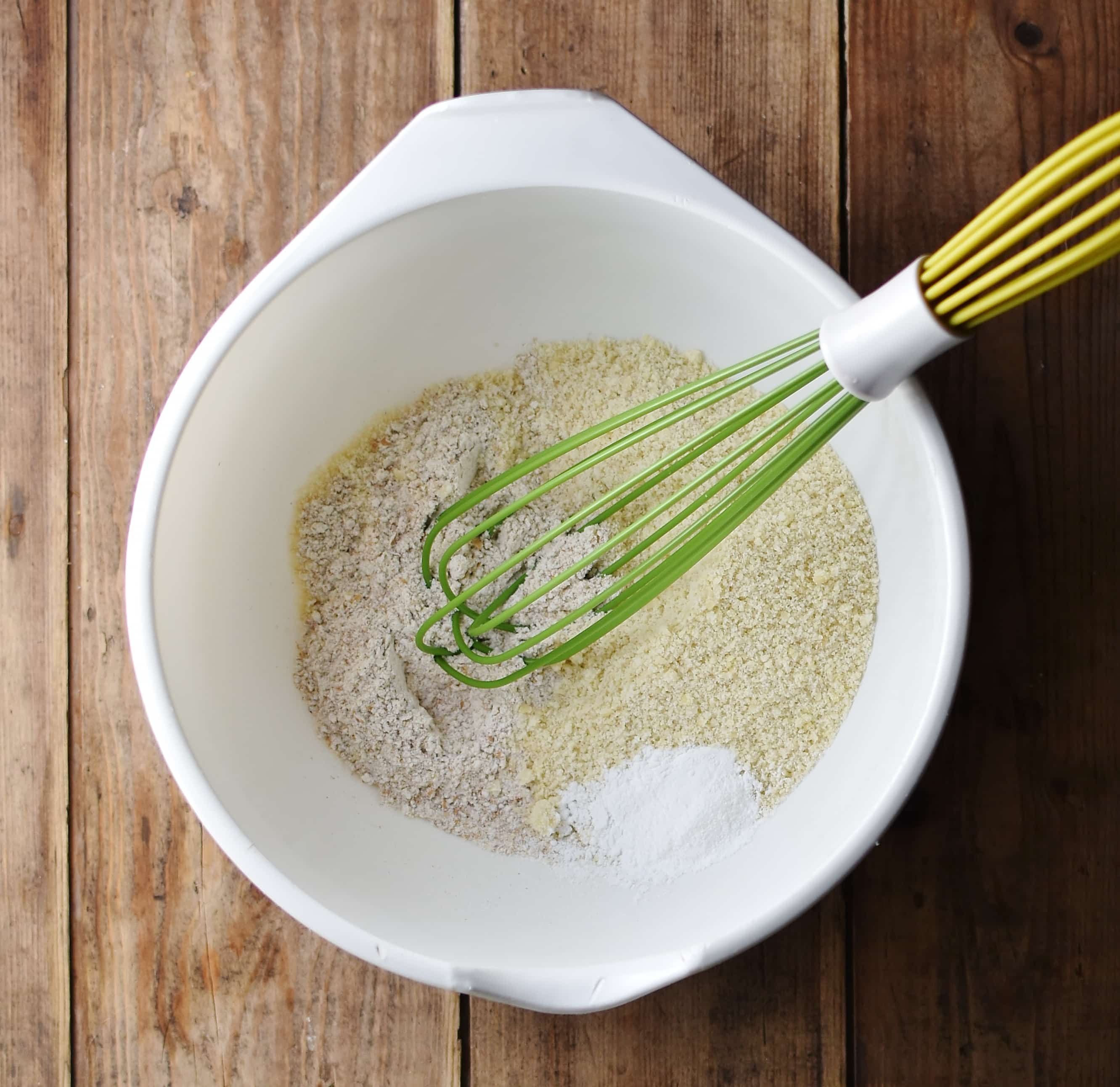 Flour mixture with green whisk on large white bowl.